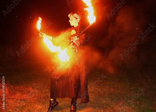Selfie portraits of a red-haired woman in a shiny gothic outfit, with horns and burning torches (double staff fire prop). Mystical fire show at night.