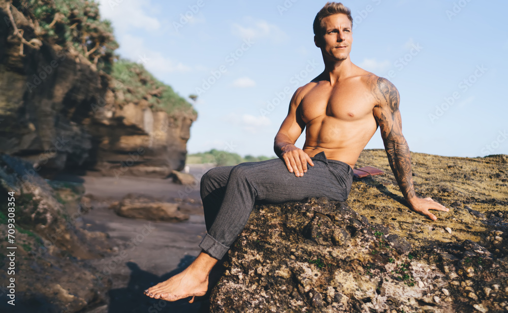 Muscular man in pants sitting on rock at seashore Muscular man in pants sitting on rock at seashore