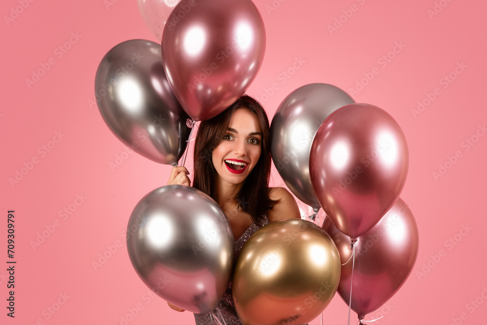 A delighted woman with dark hair and red lipstick smiles joyfully, surrounded by a cluster of metallic balloons A delighted woman with dark hair and red lipstick smiles joyfully, surrounded by a cluster of metallic balloons