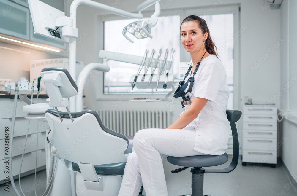 Obraz premium Portraait of sincerely smiling young dentist woman dressed white medical scrubs uniform sitting in modern dental clinic next to stomatology chair. Health care and medicare industry concept image.