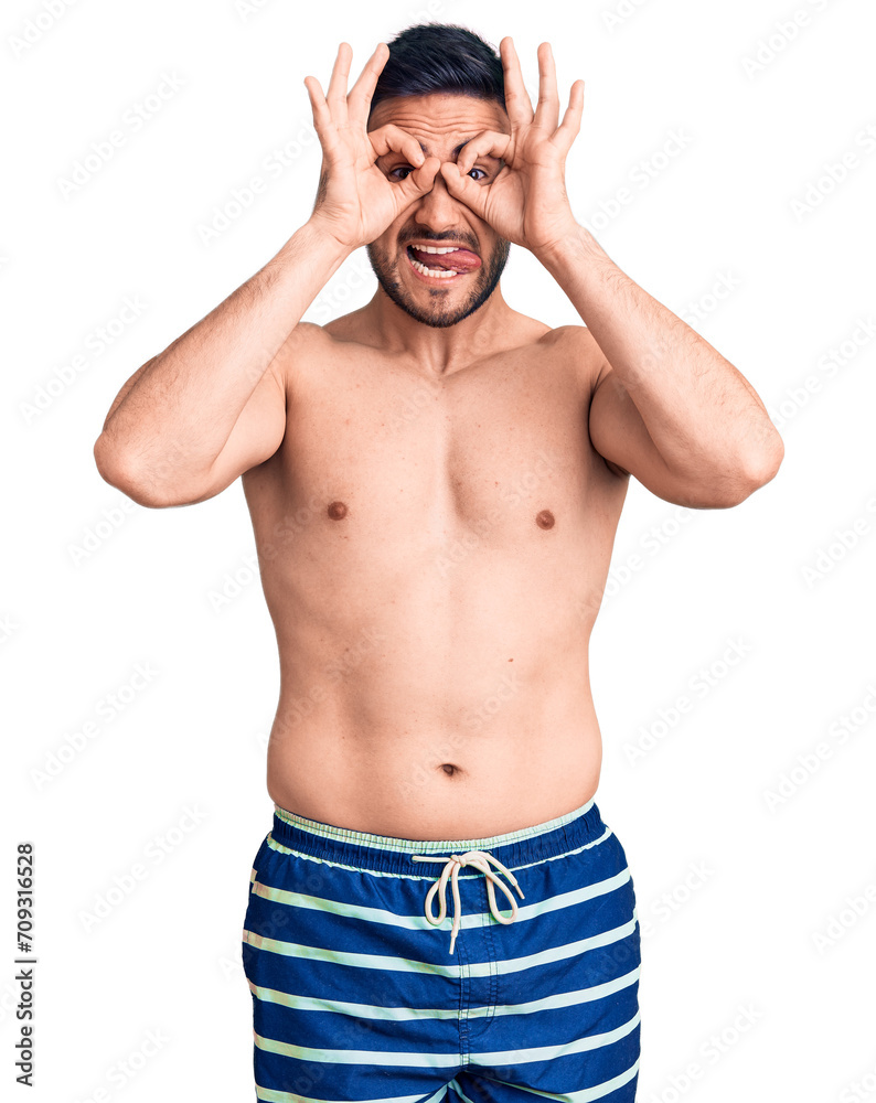 Fototapeta premium Young handsome man wearing swimwear doing ok gesture like binoculars sticking tongue out, eyes looking through fingers. crazy expression.