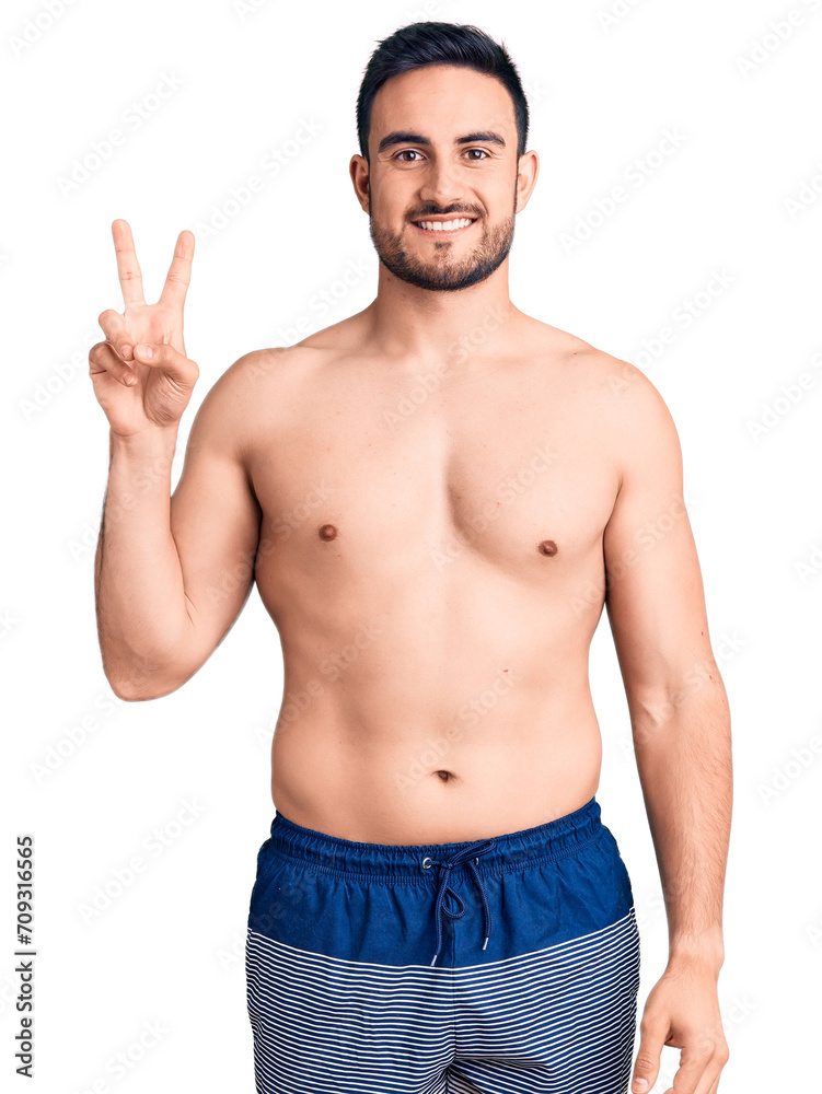 Young handsome man wearing swimwear smiling with happy face winking at the camera doing victory sign. number two. Young handsome man wearing swimwear smiling with happy face winking at the camera doing victory sign. number two.
