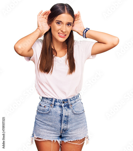 Young hispanic woman wearing casual white tshirt trying to hear both hands on ear gesture, curious for gossip. hearing problem, deaf