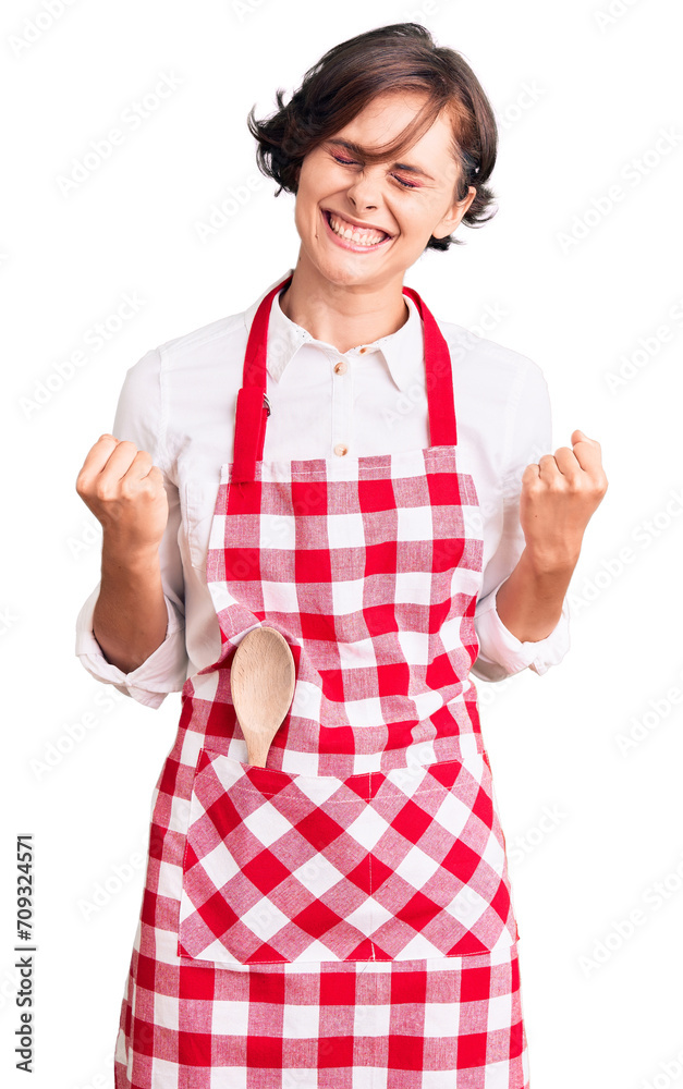Beautiful young woman with short hair wearing professional cook apron very happy and excited doing winner gesture with arms raised, smiling and screaming for success. celebration concept.