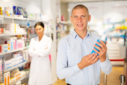 European man standing in salesroom of drugstore with bottle of sanitizer in hands. Female pharmacist standing beside shelf in background.