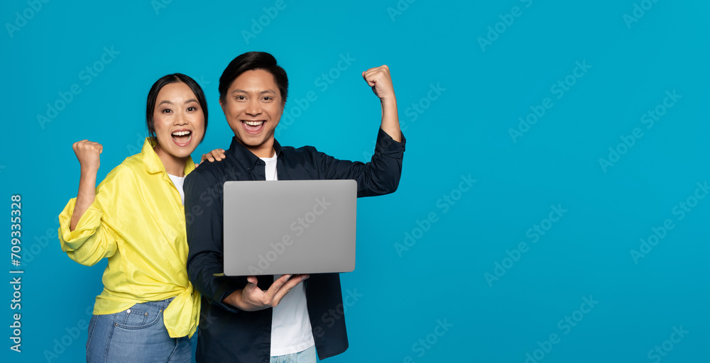 Overjoyed Asian couple triumphantly raising fists while holding a laptop, signifying success or victory