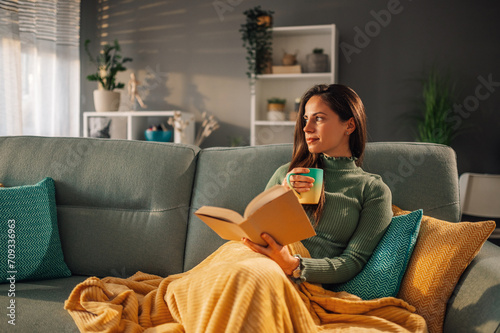 Fotografie Woman sitting on comfy couch reading a book and drinking coffee or tea