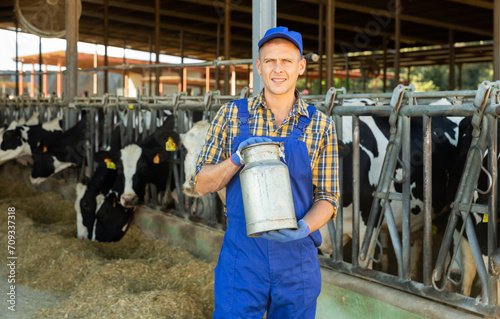 Portrait of positive dairy farm worker in blue overalls with can of milk on the background of cows in stall © JackF