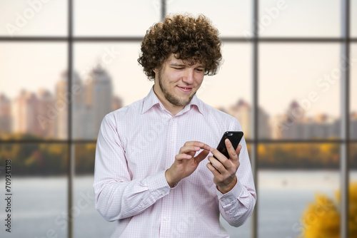 Portrait of a young smiling man with curly hair taps his smartphone. Indoor window with city river view.