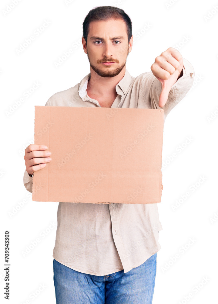 Young handsome caucasian man holding empty cardboard banner with angry face, negative sign showing dislike with thumbs down, rejection concept Young handsome caucasian man holding empty cardboard banner with angry face, negative sign showing dislike with thumbs down, rejection concept