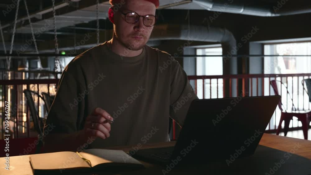 Portrait of a 30-year-old man with glasses, who is working intently in a dark room at a laptop and making notes in his notebook or notebook. A dark cafe room with ventilation pipes in the background Portrait of a 30-year-old man with glasses, who is working intently in a dark room at a laptop and making notes in his notebook or notebook. A dark cafe room with ventilation pipes in the background
