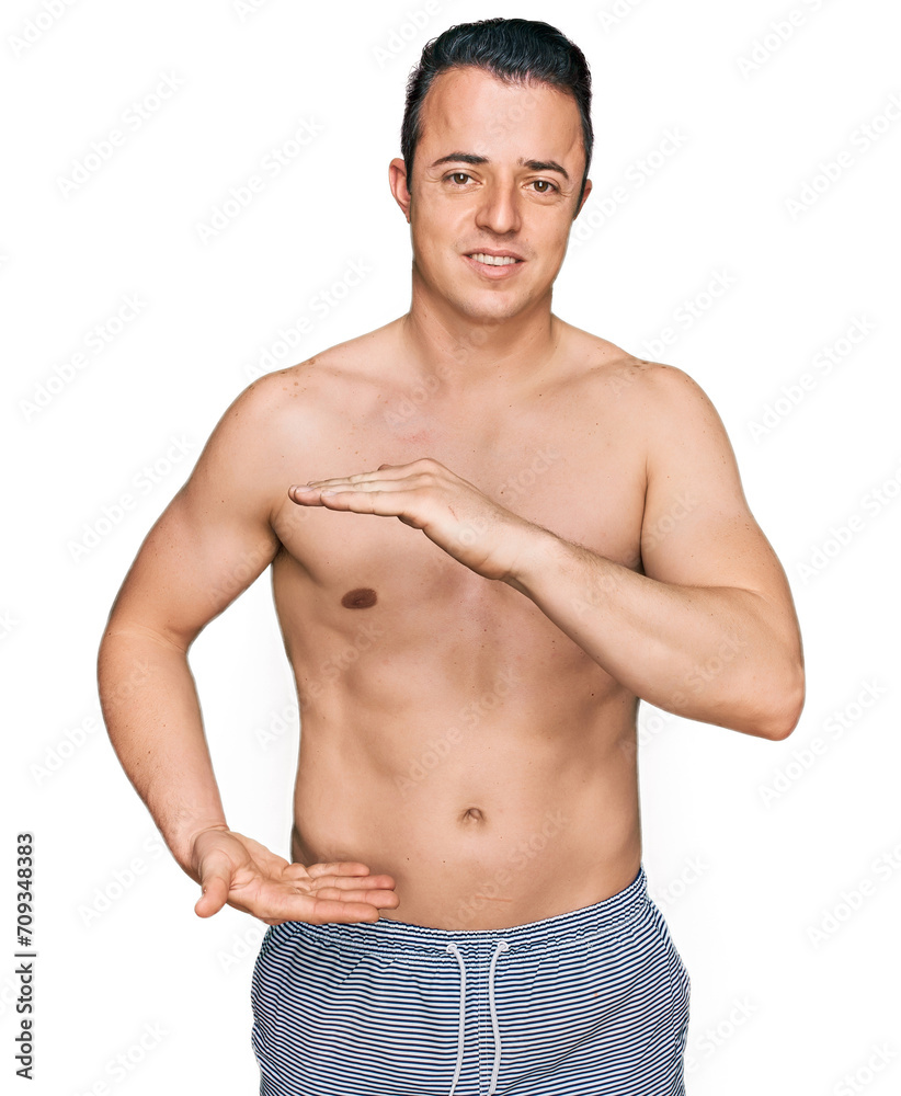Handsome young man wearing swimwear shirtless gesturing with hands showing big and large size sign, measure symbol. smiling looking at the camera. measuring concept. Handsome young man wearing swimwear shirtless gesturing with hands showing big and large size sign, measure symbol. smiling looking at the camera. measuring concept.