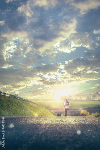 Fototapeta Naklejka Na Ścianę i Meble -  Läuferin joggt mit einem Hund auf einer Landstraße mit Gras und wunderschöner Natur und mit einem düsteren blauen Himmel und strahlender Sonne
