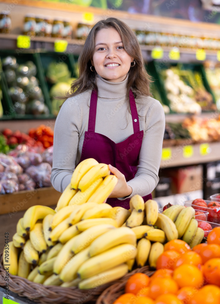 Positive young female seller holding bananas standing by counter in fruit and vegetable market