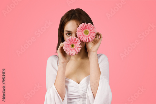 Mysterious and attractive young woman playfully holding two pink gerbera daisies over her eyes
