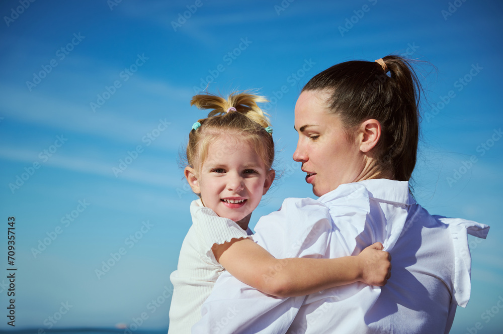 Fototapeta premium Adorable blonde Caucasian little child girl smiling looking at the camera while her loving mother carrying on the beach