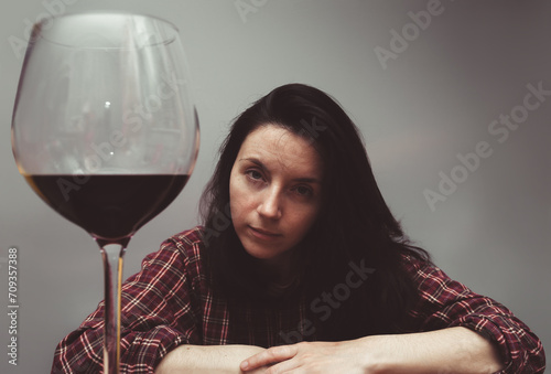 A young girl in a checkered shirt with tousled hair sits at a wooden table and looks at a large glass of red wine. Concept of alcohol abuse, headache, alcoholism, hangover, loneliness and depression. photo