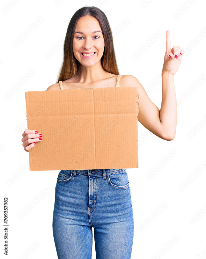 Beautiful brunette young woman holding cardboard banner with blank space surprised with an idea or question pointing finger with happy face, number one Beautiful brunette young woman holding cardboard banner with blank space surprised with an idea or question pointing finger with happy face, number one