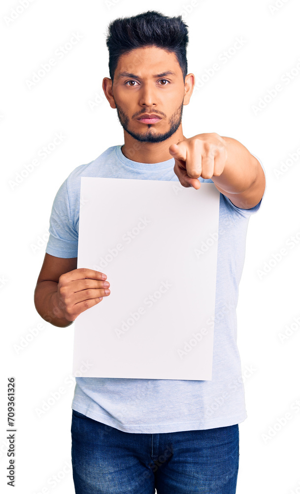 Handsome latin american young man holding cardboard banner with blank space pointing with finger to the camera and to you, confident gesture looking serious Handsome latin american young man holding cardboard banner with blank space pointing with finger to the camera and to you, confident gesture looking serious