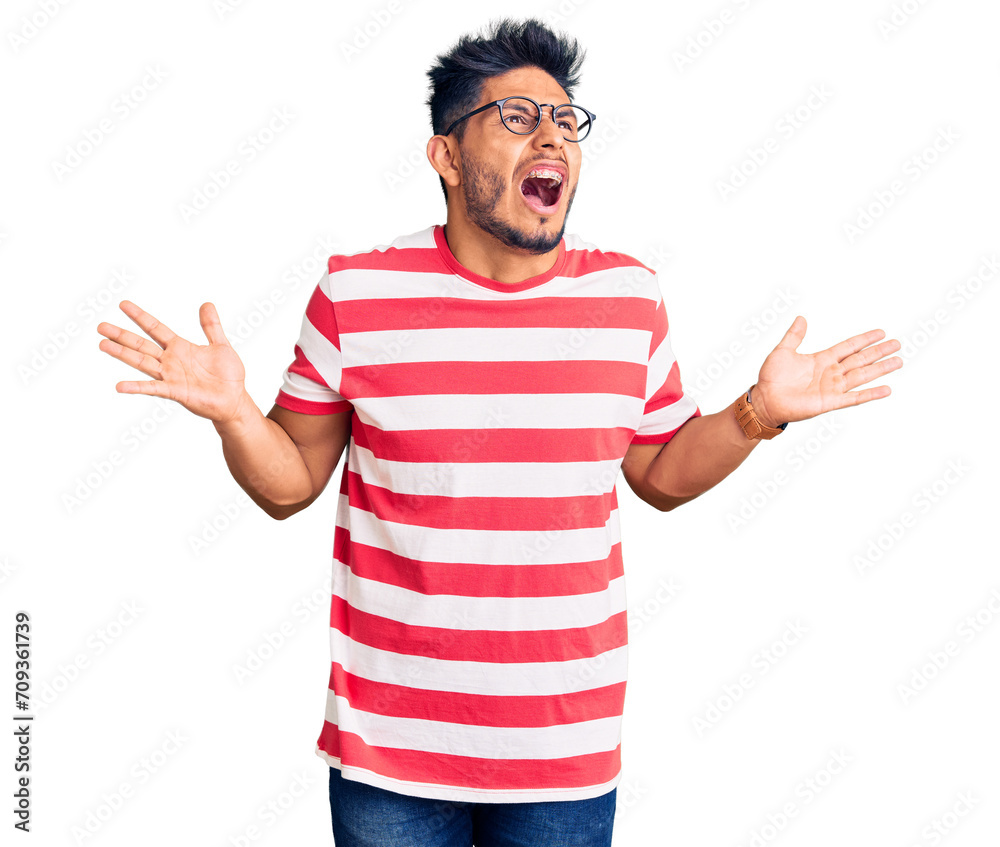 Handsome latin american young man wearing casual clothes and glasses crazy and mad shouting and yelling with aggressive expression and arms raised. frustration concept. Handsome latin american young man wearing casual clothes and glasses crazy and mad shouting and yelling with aggressive expression and arms raised. frustration concept.