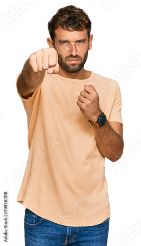 Handsome young man with beard wearing casual tshirt punching fist to fight, aggressive and angry attack, threat and violence