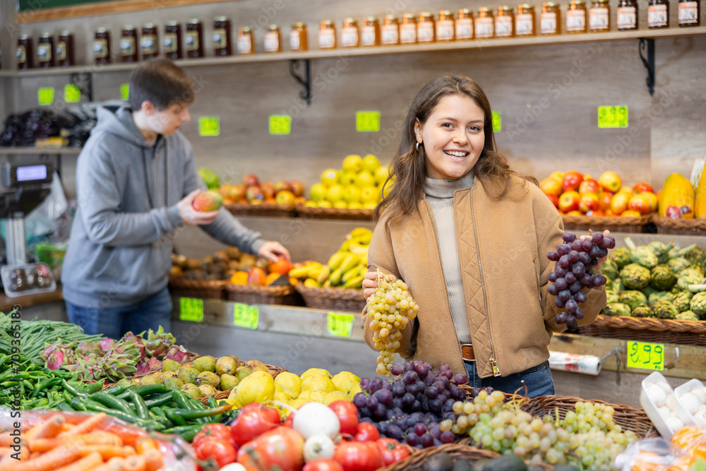 Positive young girl holding pomegranates standing at counter in large fruit and vegetable market