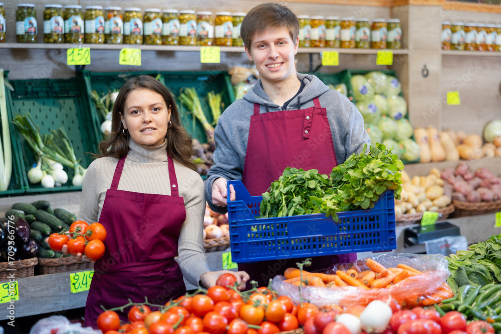 Young woman and young guy sellers laying tomatoes and celery on counter in vegetable shop