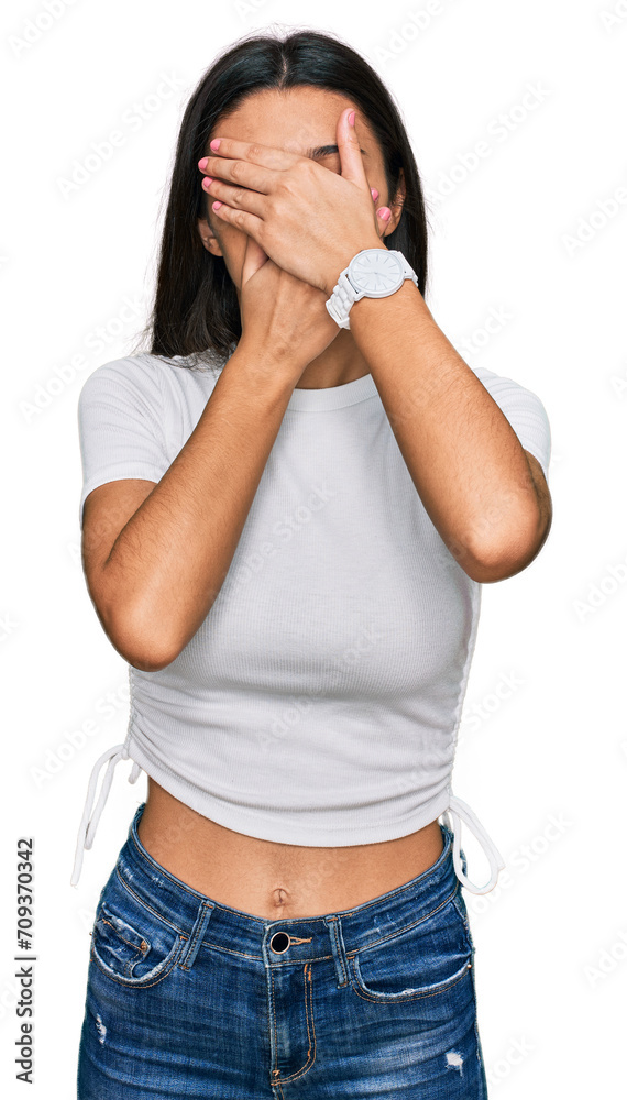 Young hispanic girl wearing casual white t shirt covering eyes and mouth with hands, surprised and shocked. hiding emotion Young hispanic girl wearing casual white t shirt covering eyes and mouth with hands, surprised and shocked. hiding emotion