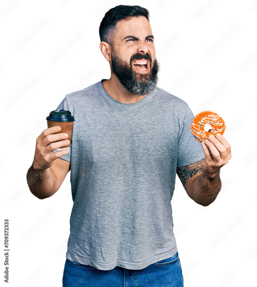 Hispanic man with beard eating doughnut and drinking coffee angry and mad screaming frustrated and furious, shouting with anger looking up.