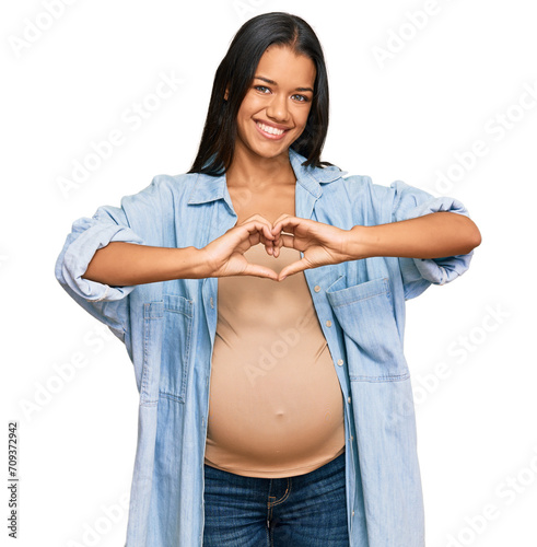 Beautiful hispanic woman expecting a baby showing pregnant belly smiling in love showing heart symbol and shape with hands. romantic concept.