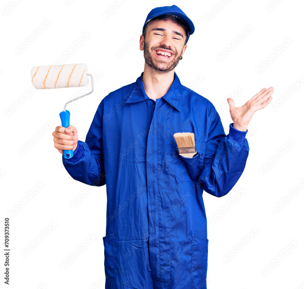 Young hispanic man wearing painter uniform holding roller celebrating victory with happy smile and winner expression with raised hands Young hispanic man wearing painter uniform holding roller celebrating victory with happy smile and winner expression with raised hands