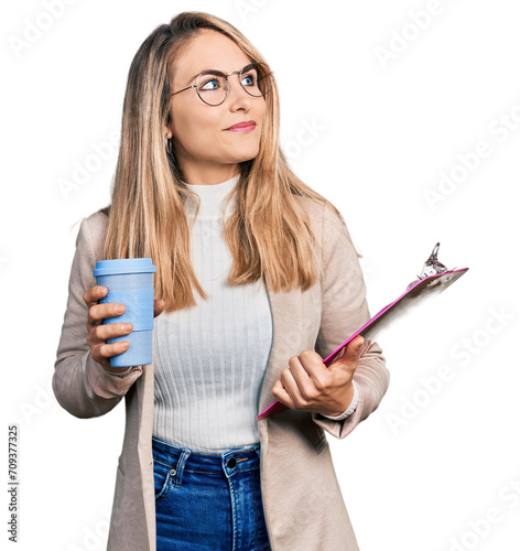 Young blonde woman wearing business style, drinking coffee and holding clipboard smiling looking to the side and staring away thinking.