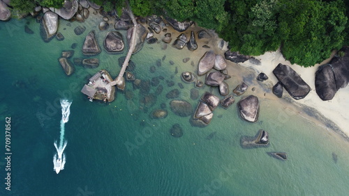 Aerial view of a hidden beach with a pier and boats, surrounded by lush greenery and large rocks, evoking peace and seclusion.