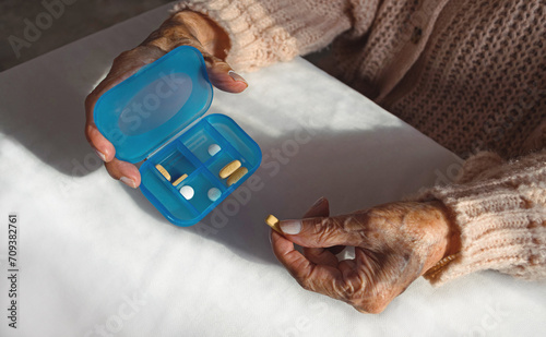 Manos de mujer anciana sosteniendo un pastillero azul y tomando una píldora o pastilla para su medicación en casa. Vista en primer plano o closeup. 