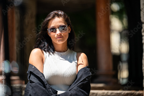 Portrait of an attractive young woman wearing summer garments and sunnglasses in a city landscape with blurred background on a bright sunny day
