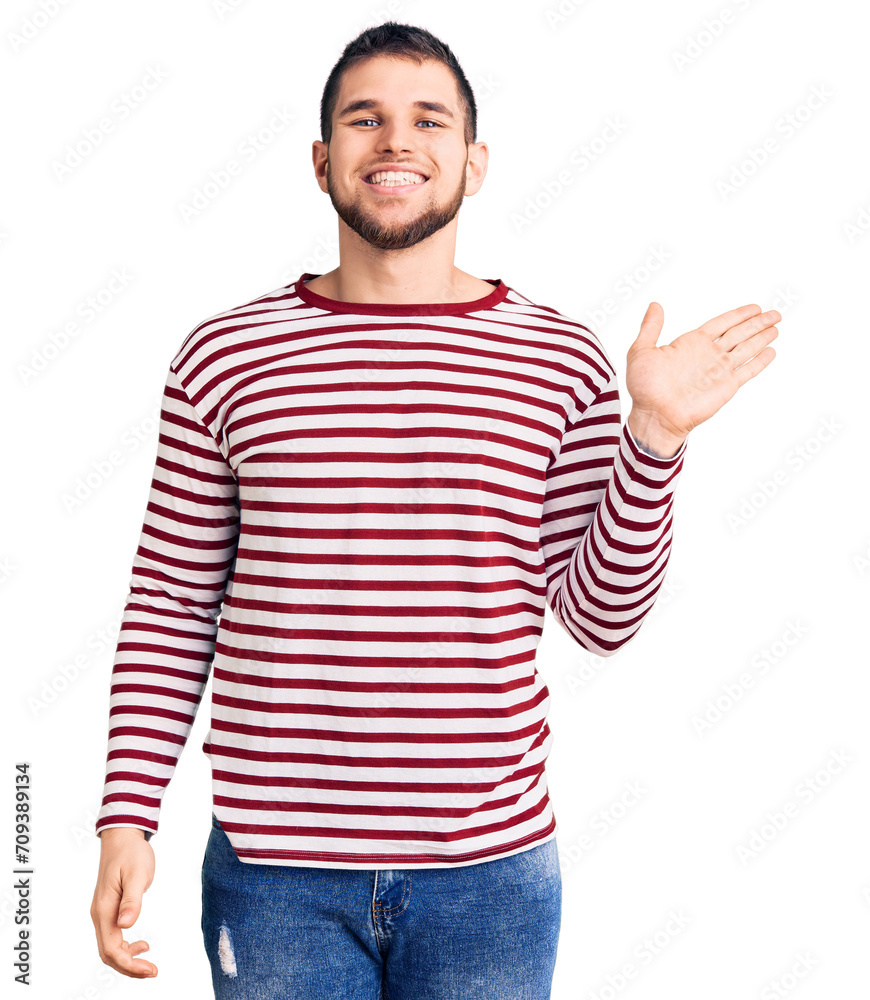 Young handsome man wearing striped sweater smiling cheerful presenting and pointing with palm of hand looking at the camera. Young handsome man wearing striped sweater smiling cheerful presenting and pointing with palm of hand looking at the camera.