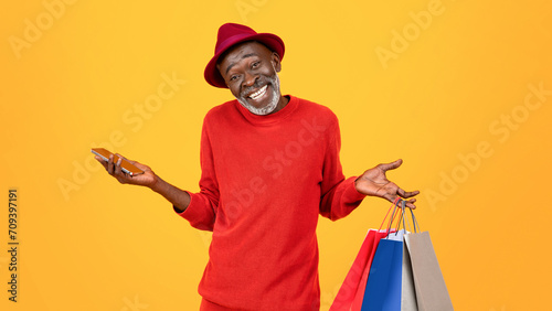 Smiling confused old black man shopaholic in hat, shrugs shoulders with many bags photo