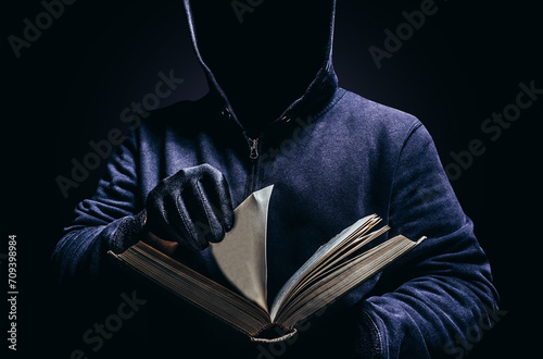 Fotografie Photo of scary shaded hooded man holding book and turning the pages on dark background
