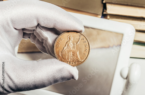 Photo of a person's male hand in white gloves holding a british one penny 1945 coin on stack of books and magnifying glass background. Numismatics hobby concept.