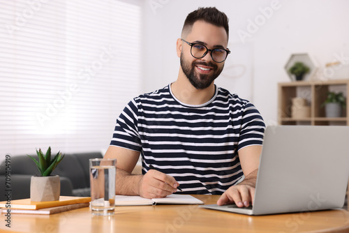 Young man writing in notebook while working on laptop at wooden table indoors