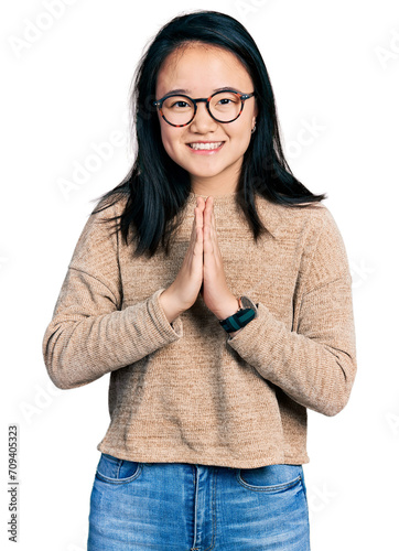 Young chinese woman wearing casual sweater and glasses praying with hands together asking for forgiveness smiling confident.