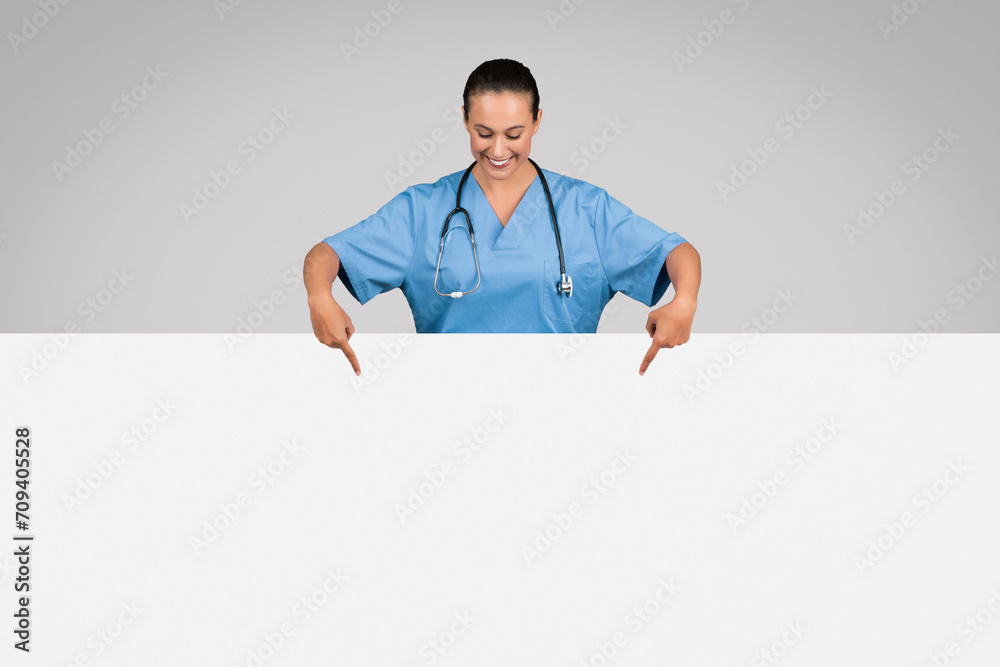 Lady nurse in blue medical uniform pointing down at white placard board against grey background, banner Lady nurse in blue medical uniform pointing down at white placard board against grey background, banner