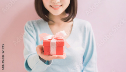 リボンがかかった赤い小さなプレゼントの箱を、胸の前に差し出す若い女性の上半身のAI画像