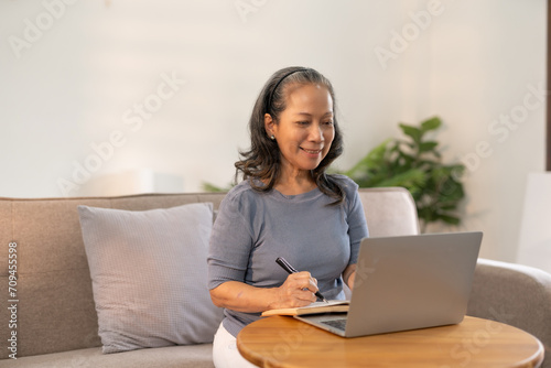 Senior asian woman sitting on sofa using laptop computer, working, chatting, social media internet at home. © Premreuthai