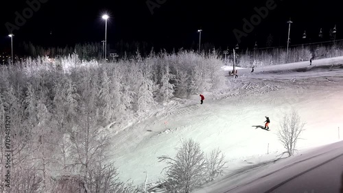 Aerial shot of snowboarders moving down the ski-run at night. Snowfields and forest in the distance. Doing winter sports in the evening