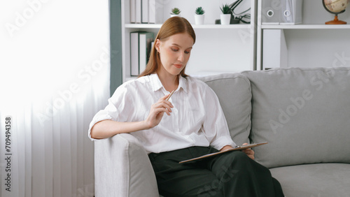 Professional female psychologist with clipboard portrait sitting on arm chair in psychiatrist office or therapy room. Friendly and prim mental healthcare counselor and therapist.