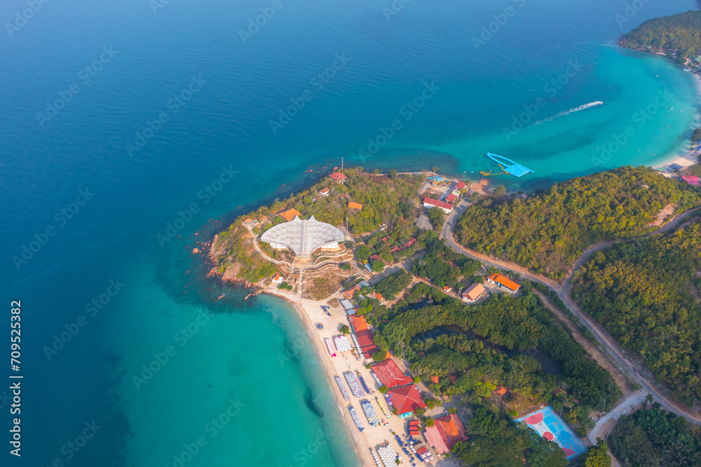 Tropical island with a sandy bay and pier and azure beach village settlement surrounded by exotic green forests trees, aerial view. Resort beach with umbrellas and sun loungers for tourists in hotels