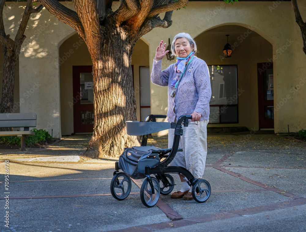 An elderly woman with a facemask under her chin waving to the camera, walking outdoors with a mobility walker in a retirement village.