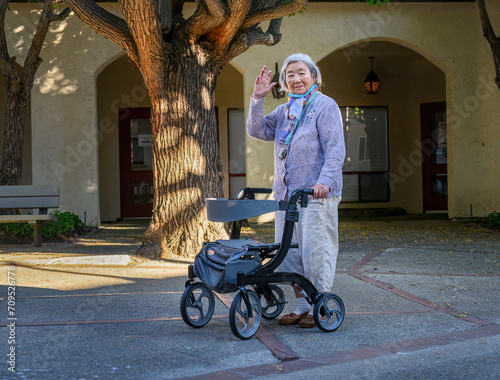 Obraz na płótnie An elderly woman with a facemask under her chin waving to the camera, walking outdoors with a mobility walker in a retirement village. w salonie