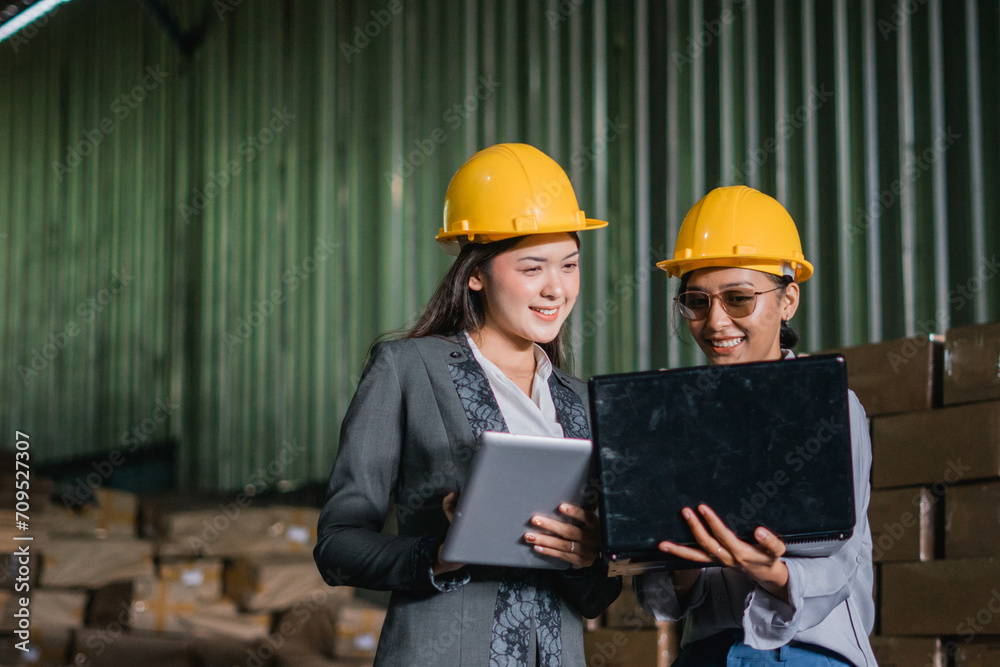 two young women factory workers wear safety helmets while using a tablet and laptop computers at the factory two young women factory workers wear safety helmets while using a tablet and laptop computers at the factory